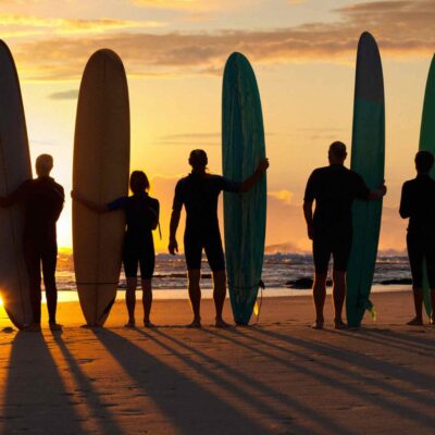 Silhouettes of six surfers holding surfboards standing on a beach at sunset.