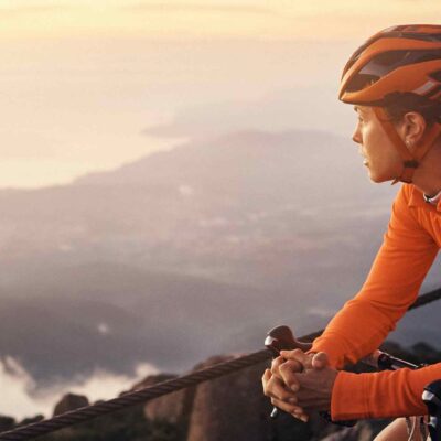 A cyclist in an orange jersey and helmet rests on a guardrail, gazing at a misty sunrise over a coastal landscape.
