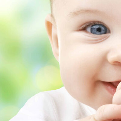 Close-up of a smiling baby with bright blue eyes, sucking on their hand, against a soft green and blue blurred background.