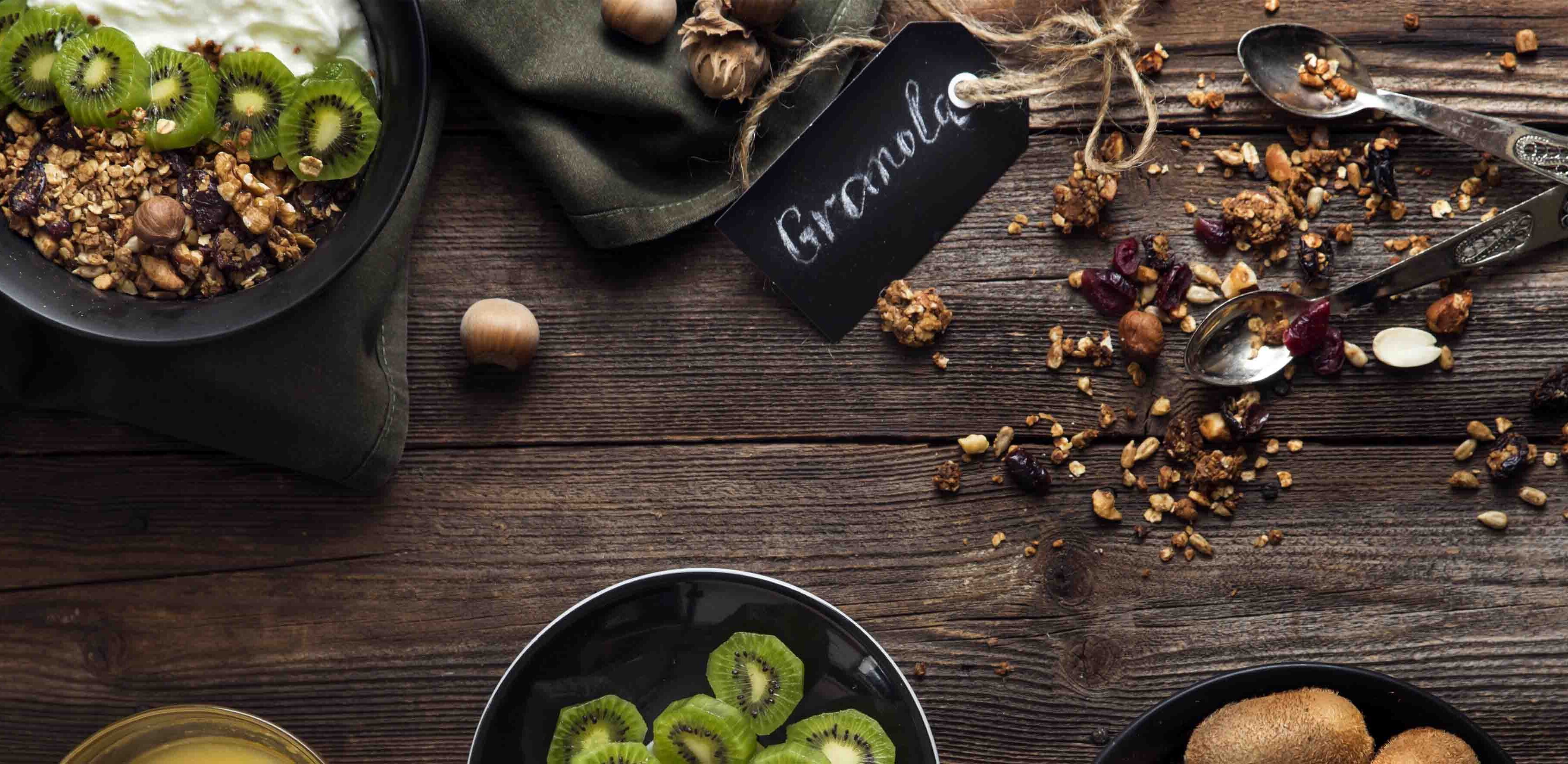 Healthy breakfast spread on a wooden table, featuring bowls of granola with yogurt and kiwi slices, nuts, and dried fruits.