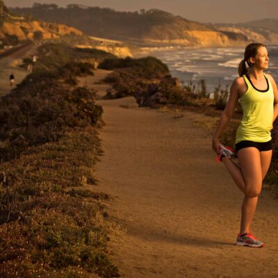 A woman stretching on a coastal trail at sunset, with cliffs and the ocean in the background.