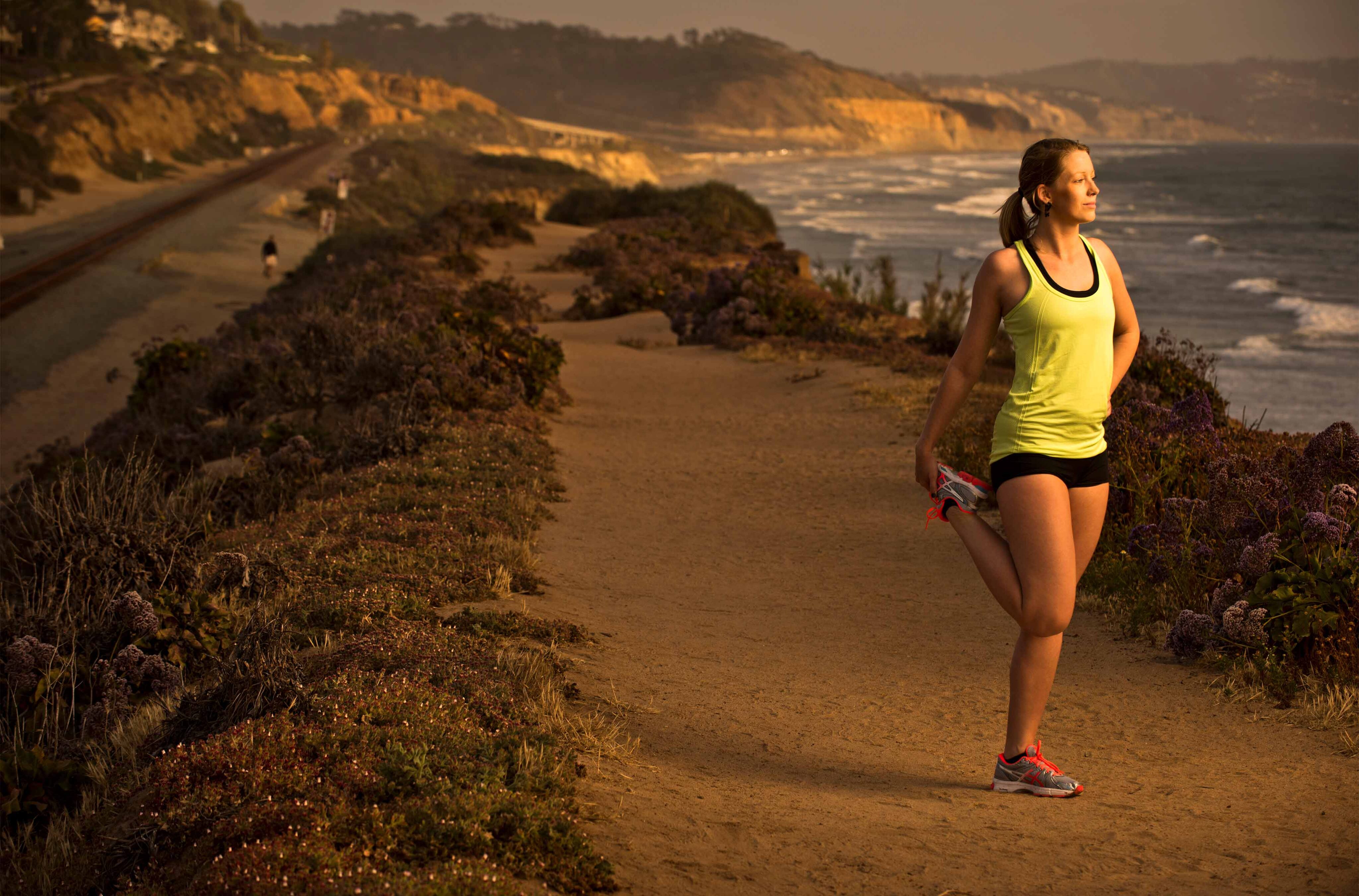 A woman stretching on a coastal trail at sunset, with cliffs and the ocean in the background.