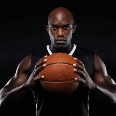 A focused male basketball player holding a basketball, wearing a black tank top against a dark background.