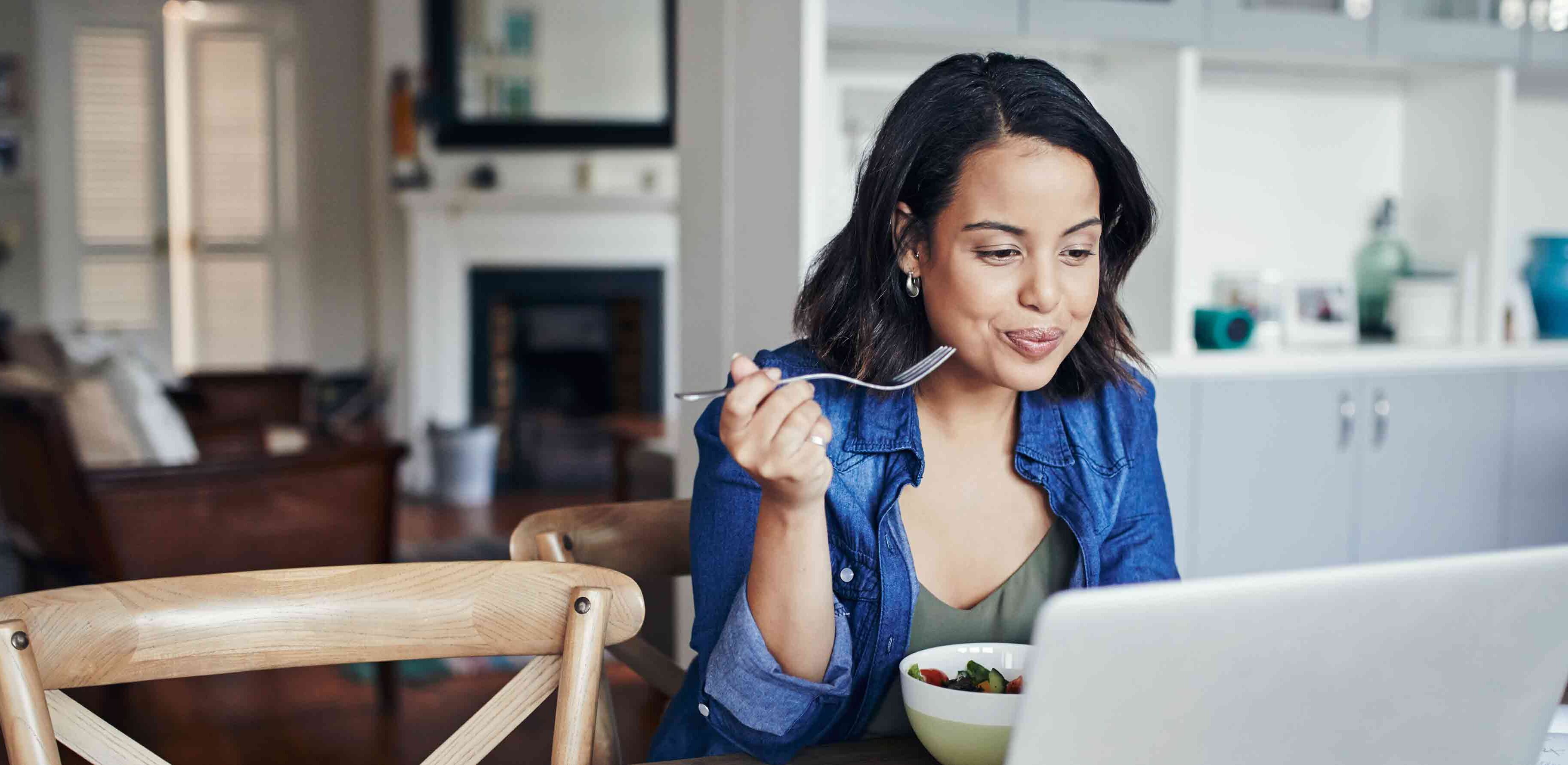 A woman eating a salad while working on a laptop in a stylish kitchen.