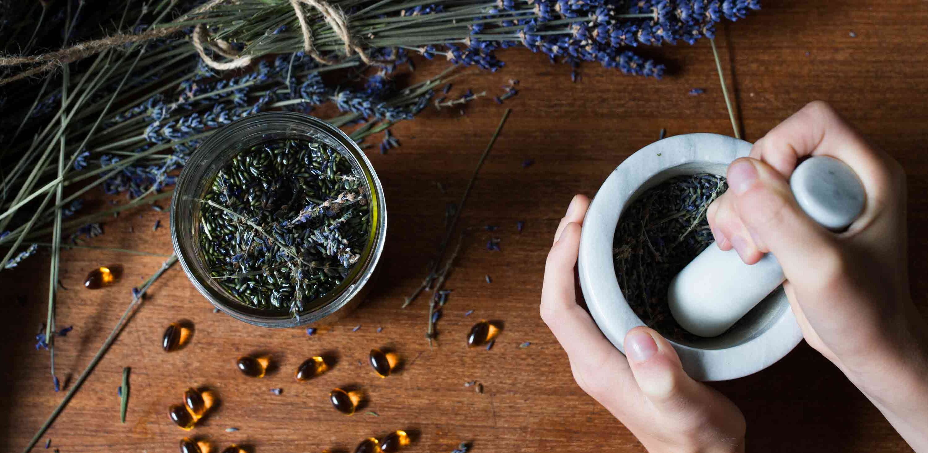 Hands grinding dried herbs in a mortar and pestle on a wooden table, with scattered lavender and small amber beads around.