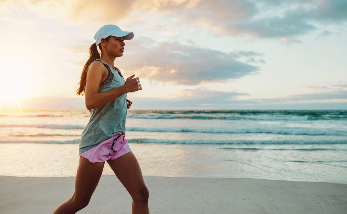 Woman jogging on a beach at sunset, wearing a cap, tank top, and pink shorts, with the ocean in the background.