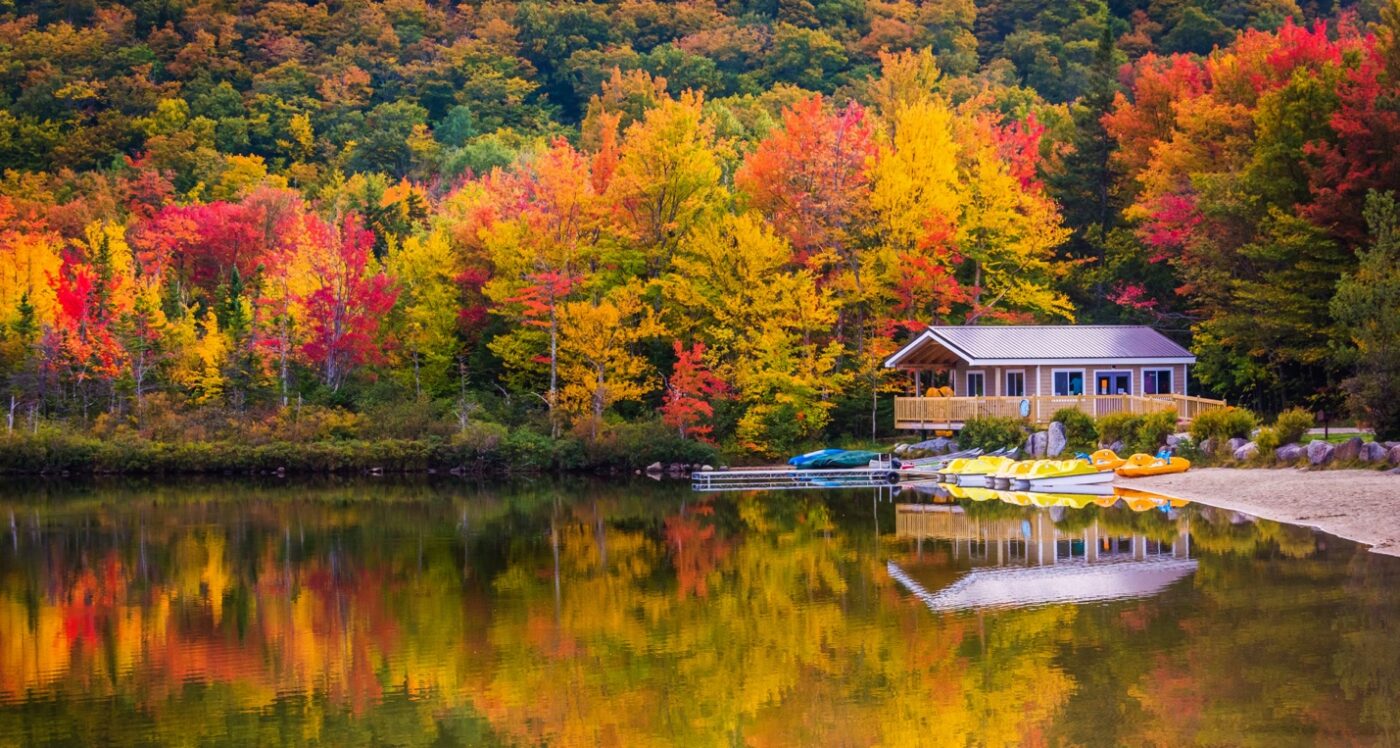 Autumn-colored trees reflecting in a serene lake with a small house and docked kayaks on the shore.