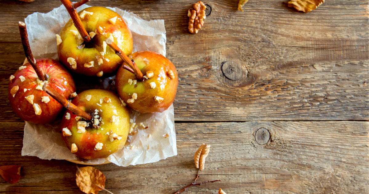 Caramel apples with nuts and sticks on a rustic wooden table, surrounded by scattered autumn leaves.