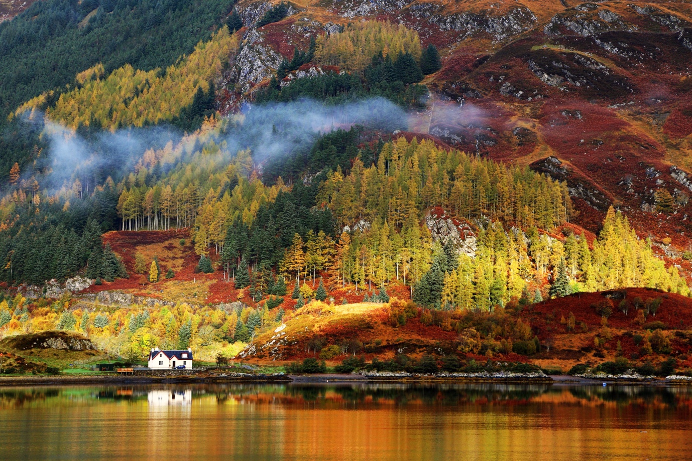 Autumn landscape with colorful trees surrounding a white house by a lake, with mist rising from the forested hills.