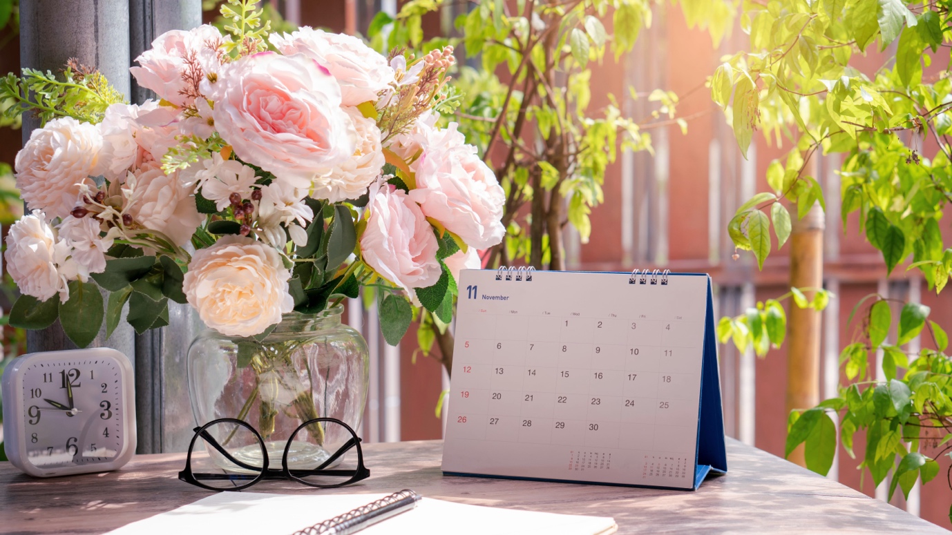 A serene workspace with a vase of pink roses, a calendar, glasses, and notebook on a desk beside a window with leafy plants.