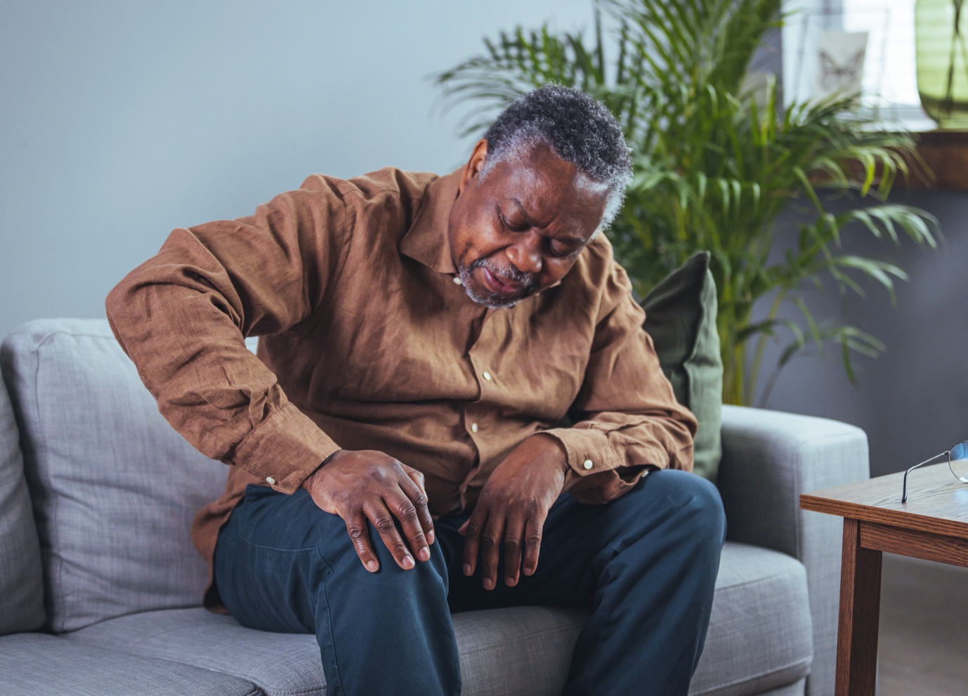 An elderly black man sitting on a sofa, looking down with a pained expression, possibly in distress or deep in thought.