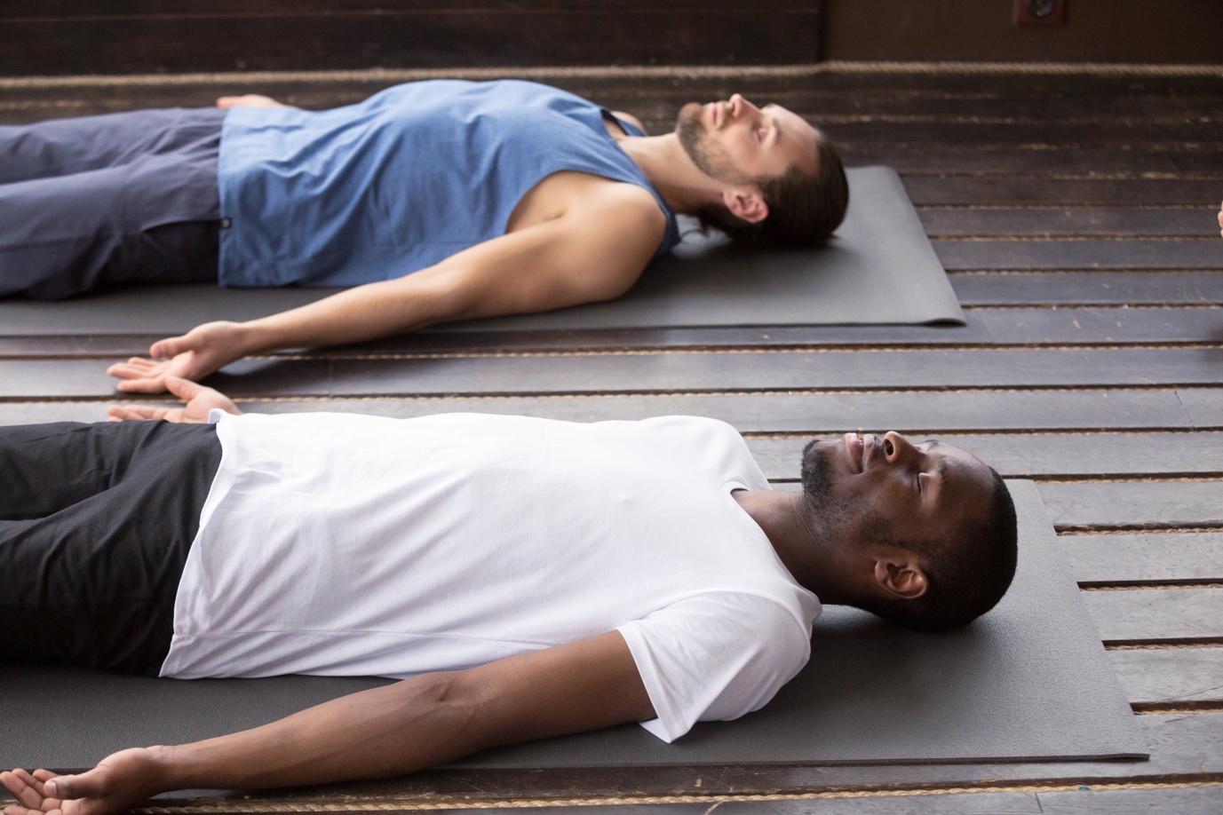 Two men lying on yoga mats in savasana pose, one in a white shirt, the other in a blue tank top, on a wooden deck.