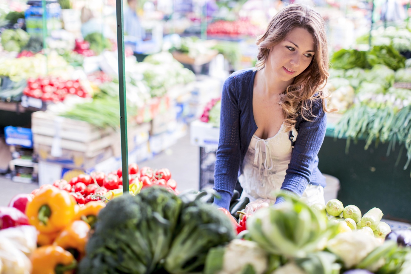A woman selects vegetables at a vibrant outdoor market, surrounded by fresh produce in varied colors.