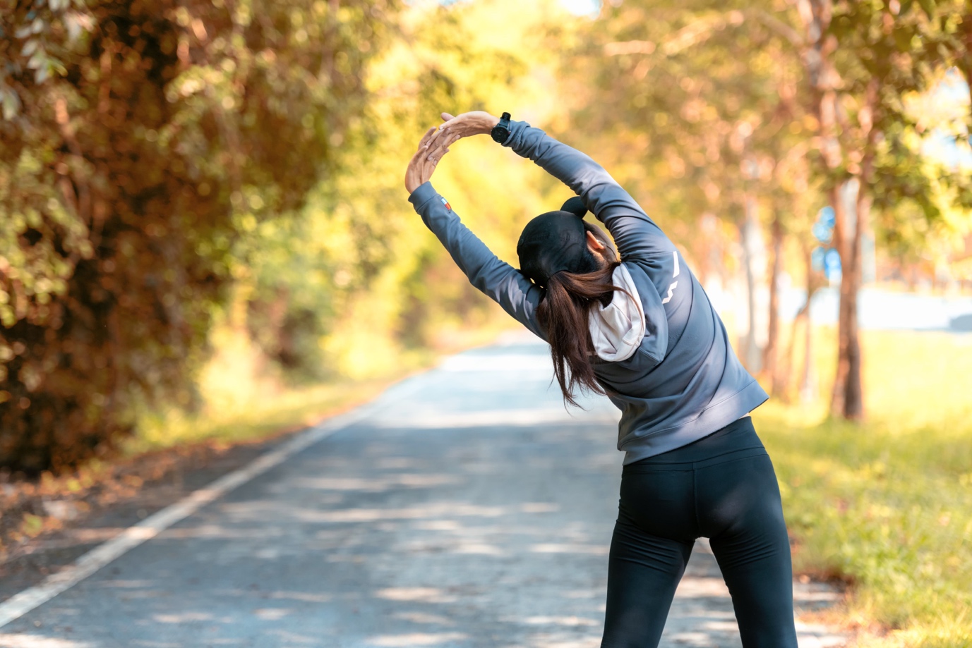 A woman stretches her arms overhead on a sunlit path lined with trees, wearing sportswear and a ponytail.