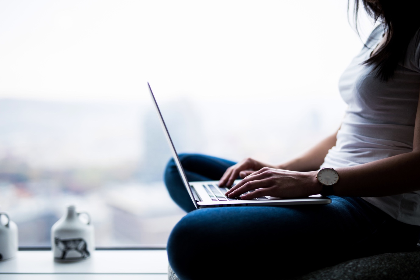 A woman sits by a window, using a laptop on her lap, with a blurred cityscape in the background. she is wearing a white watch.