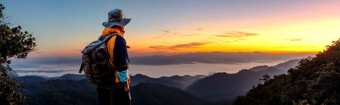 Hiker wearing a hat and backpack admiring a sunrise over a mountainous landscape with mist-covered valleys.