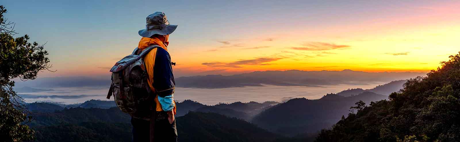 Hiker wearing a hat and backpack admiring a sunrise over a mountainous landscape with mist-covered valleys.