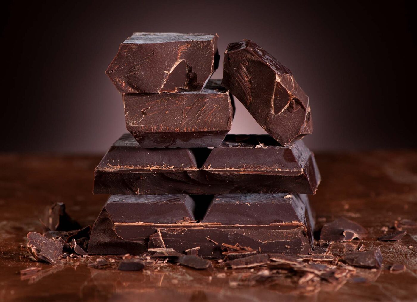 Stack of dark chocolate pieces with shavings on a brown surface.