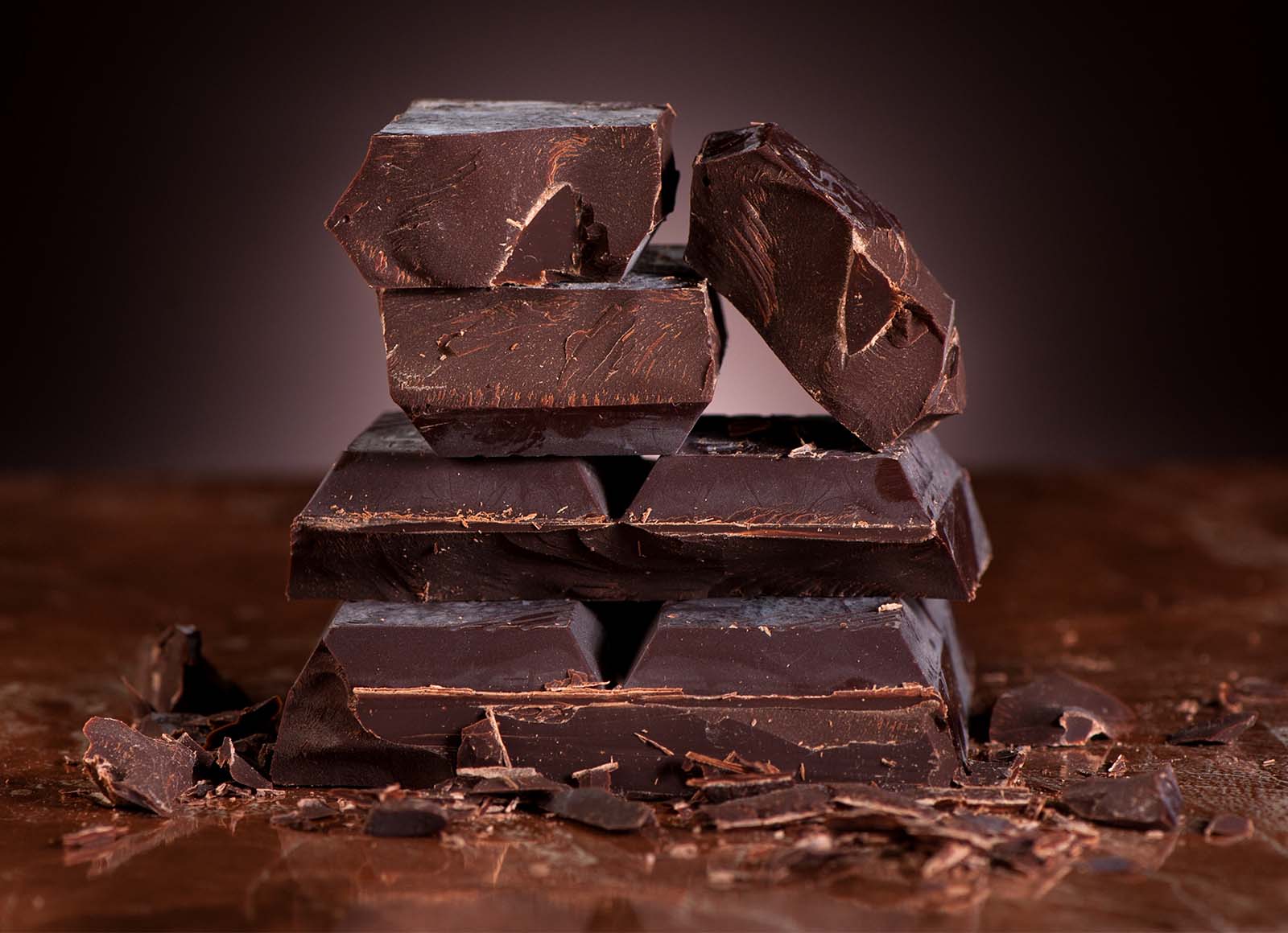 Stack of dark chocolate pieces with shavings on a brown surface.