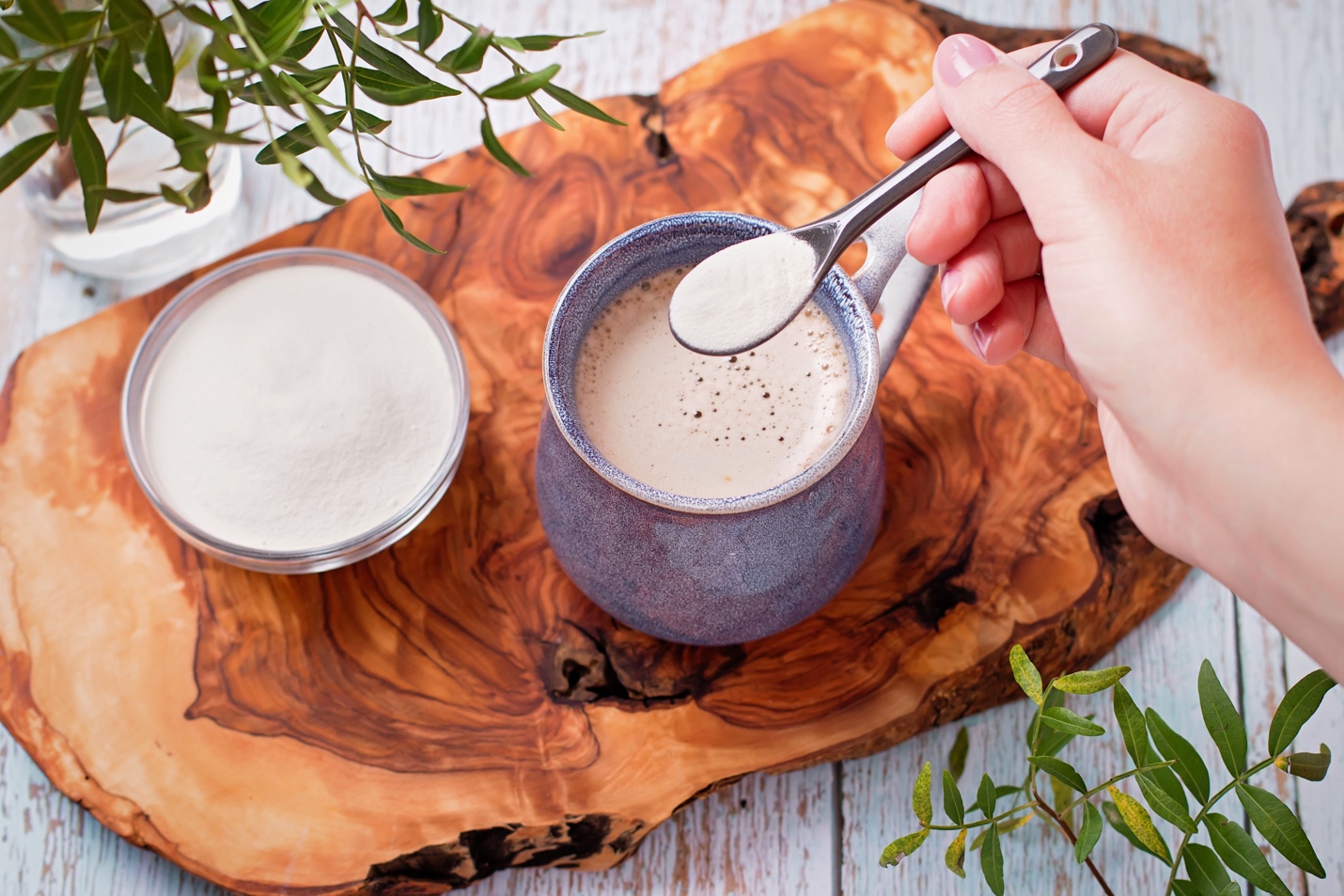 A hand sprinkling collagen powder into a cup of coffee on a wooden slab, with a bowl of collagen powder nearby.