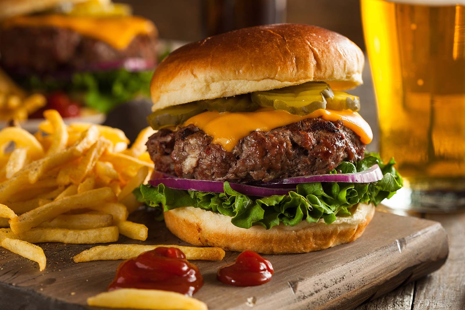 Cheeseburger with lettuce, onions, and two patties on a board, surrounded by fries and condiments, with a glass of beer in the background.