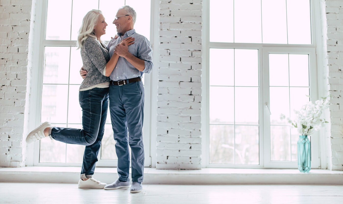 A joyful elderly couple dances in a bright room with large windows, the woman lifted slightly off the floor as they embrace.