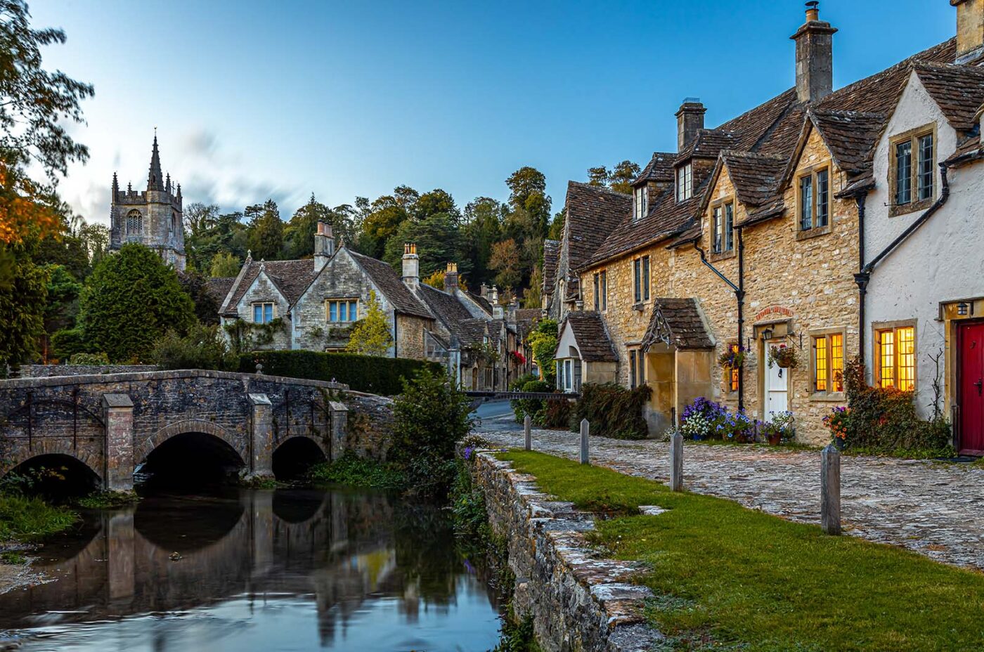 Quaint english village scene with stone bridge over a calm river, lined by traditional cottages and a church spire in the background at dusk.