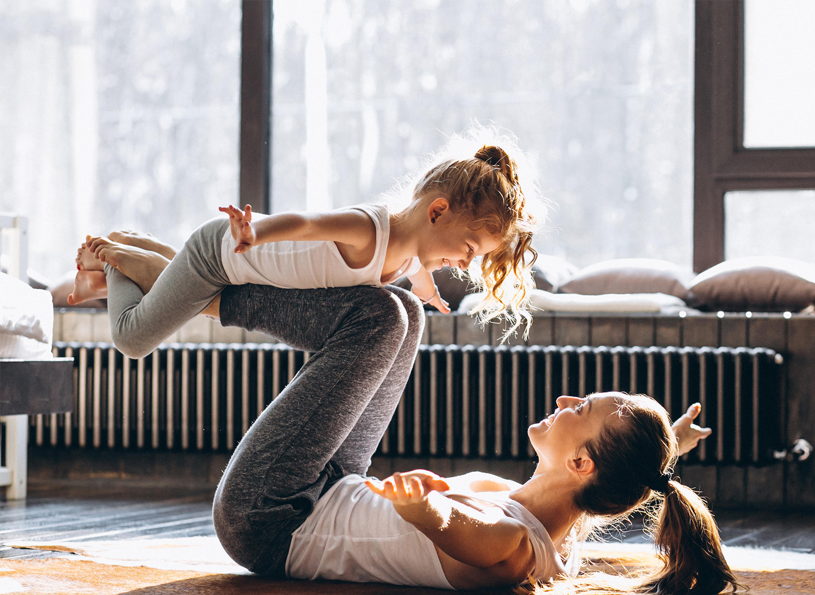 A woman and a young girl practice yoga together in a sunlit room, performing a playful airplane pose.