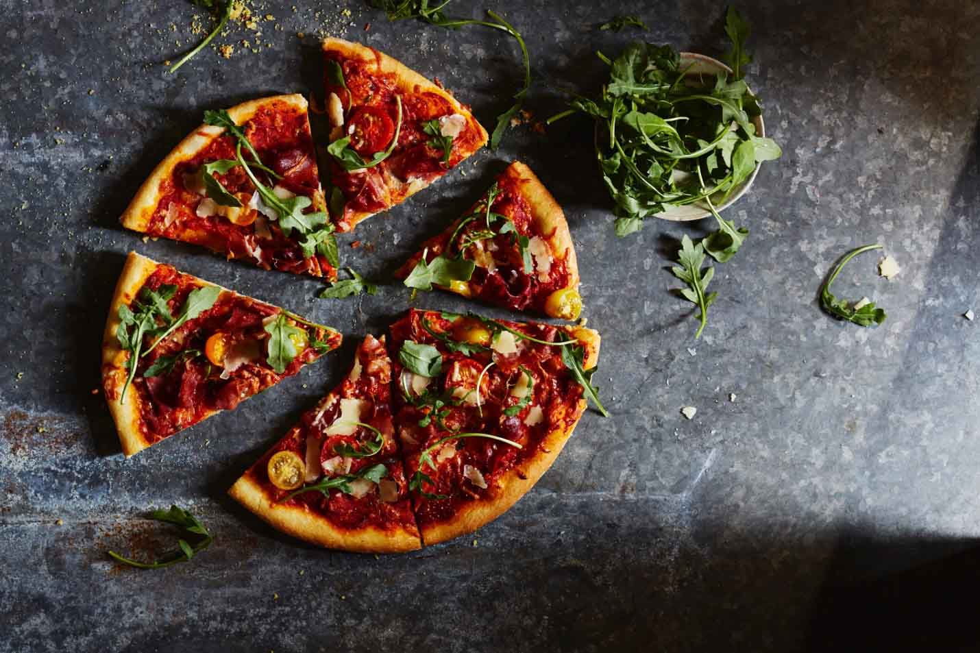 Sliced pizza with toppings of arugula, cheese, and slices of meat on a dark, textured surface.