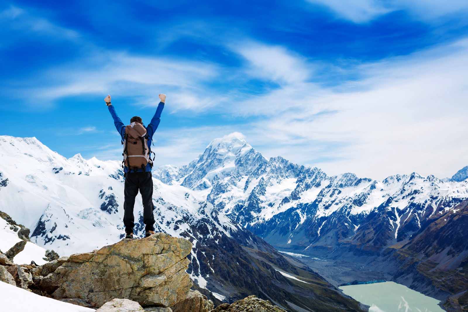 Man raising arms in triumph on a mountaintop, overlooking snowy peaks and a glacier.