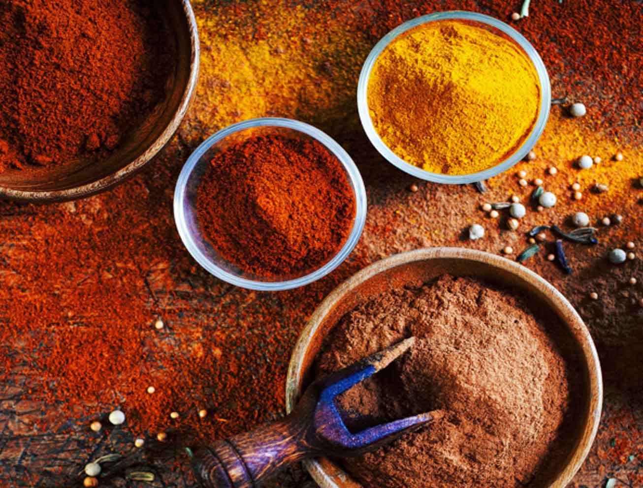 Various spices including chili powder, turmeric, and cumin in bowls on a rustic surface sprinkled with seeds and peppercorns.
