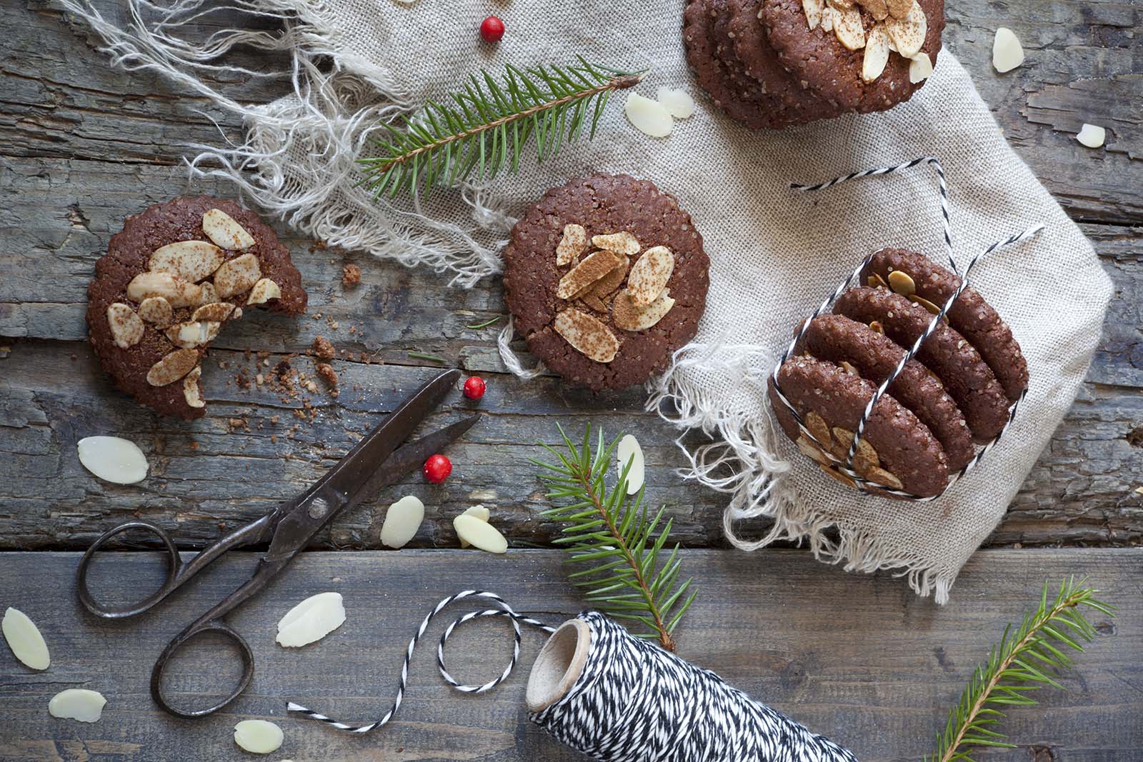 Rustic wooden table with chocolate almond cookies, vintage scissors, twine, and scattered almond slices, decorated with pine branches and red berries.