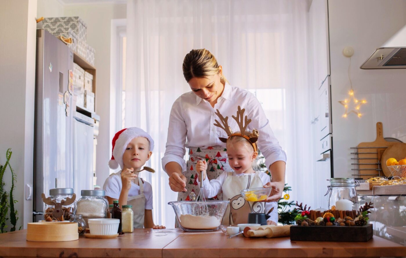 A woman and two children in festive hats baking cookies in a kitchen decorated for christmas.